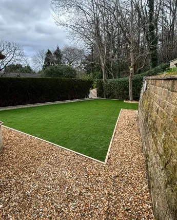 Finished artificial lawn with crisp edging and gravel borders beside a stone boundary wall