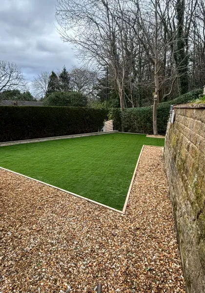 Finished artificial lawn with crisp edging and gravel borders beside a stone boundary wall