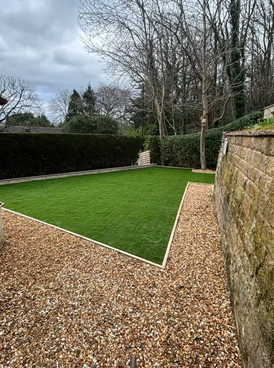 Finished artificial lawn with crisp edging and gravel borders beside a stone boundary wall